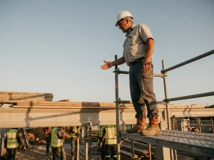 Bouwvoorman op steiger geeft instructies aan ploeg beneden, witte helm en stalen neuzen, gouden-uurlicht over beton en hout.