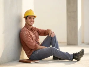 Jonge bouwvakker met helm leunt ontspannen tegen betonnen muur, notitieboek en smartphone naast werkschoenen.