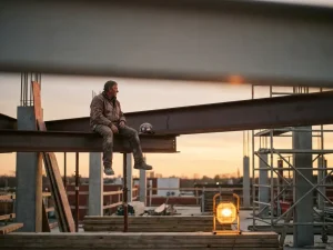 Bouwvakker rust op stalen balk bij steigers in warm middaglicht, helm naast zich, betonnen bouwplaats op de achtergrond.