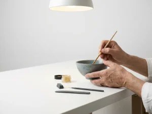 Handen die een gebarsten keramische kom restaureren met gouden lak, kintsugi-techniek op wit werkoppervlak.