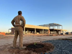 Nieuwkomer in werkkleding bij bouwplaats kijkt vooruit naar half gebouwd gebouw, klein groen plantje in verse aarde aan hun voeten.